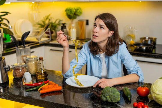 Young woman feels sad and frustrated holding measuring tape in home kitchen thinking about strict weight control. Adult girl questions progress and fights cravings using willpower for healthy eating.