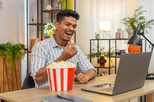 Indian man at home watches comedy film on laptop eats popcorn laughs and enjoys funny scene. Arabian handsome guy at desk smiles takes bite from backet looks at screen feels joy relax and happy mood