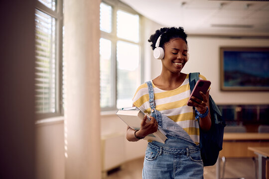 Happy black student using her smart phone while listening music over headphones at university.