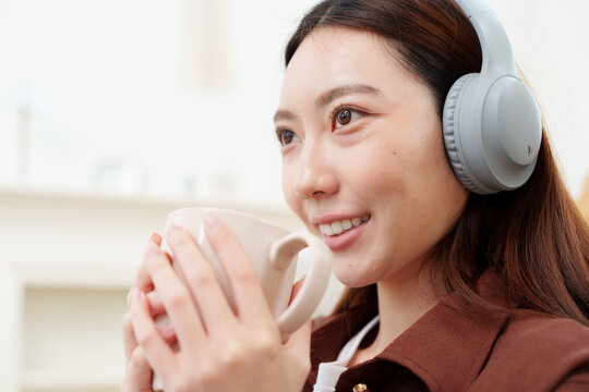Close up of young, beautiful Japanese woman in brown shirt with headphones looking ahead, holding warm coffee or tea mug with both hands, smelling with gentle smile in cozy living room at home