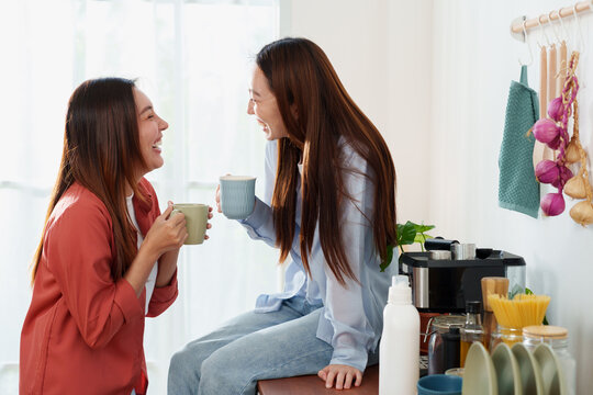 Two young Asian woman relaxing, smiling, laughing together with coffee mug in hands, one standing and the other sitting on counter in cozy kitchen at home