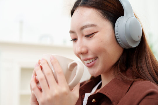Close up of young, beautiful Japanese woman in brown shirt with headphones holding and looking at coffee or tea mug with both hands, smelling with gentle smile in cozy living room at home