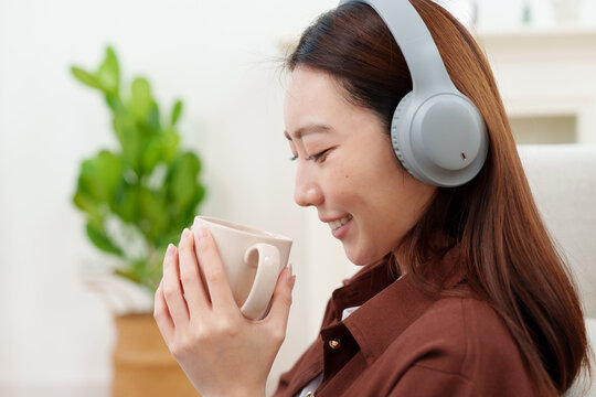 Close up of young, beautiful Japanese woman in brown shirt with headphones holding and looking at coffee or tea mug with both hands, smelling with gentle smile in cozy living room at home
