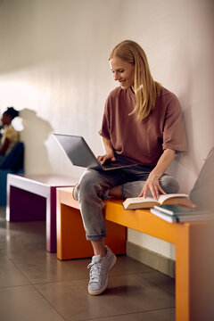 Happy college student surfing the net on laptop while learning in hallway.