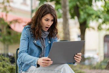 Fototapeta premium Young woman finding laptop error in city street park with shocked stressed mood at work. Girl tourist covering mouth outdoors after spotting mistake on computer during freelance work break near trees.