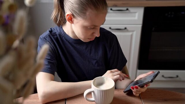 Young person at kitchen table tapping smartphone beside white mug in warm domestic home interior with blurred cabinets, focused expression, soft daylight, and shallow depth of field
