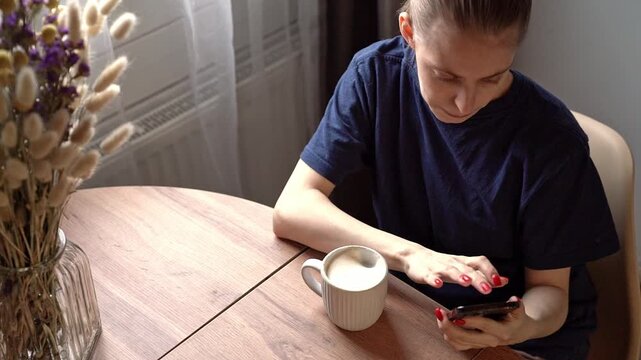 Woman using smartphone beside coffee mug at wooden dining table, with hand swiping across screen, soft daylight, dried flowers, calm home interior, top angled view