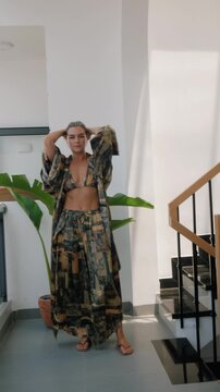 Woman modeling flowing patterned resort wear with bikini top and wide leg pants in bright modern interior.