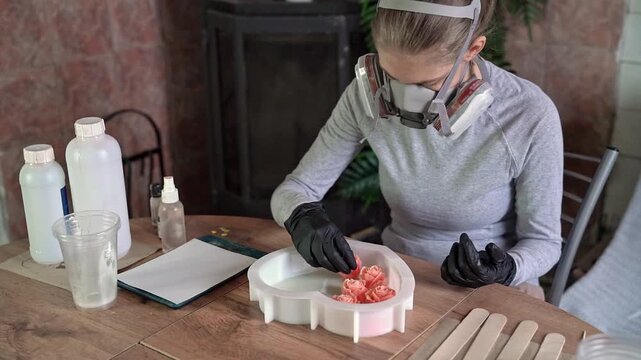 Craftsperson placing pink resin roses into silicone mold during indoor workshop with gloved hands and protective respirator, creating glossy floral craft piece on wooden table