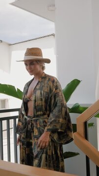Woman modeling patterned resort wear with bikini top, flowing robe and straw hat in sunlit modern interior.