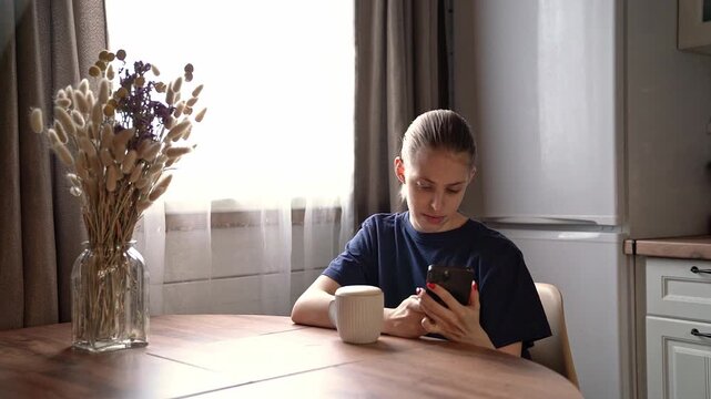 Young woman sitting at wooden dining table in bright home kitchen, looking down at smartphone with calm focus beside ceramic mug and dried flowers near sunlit window