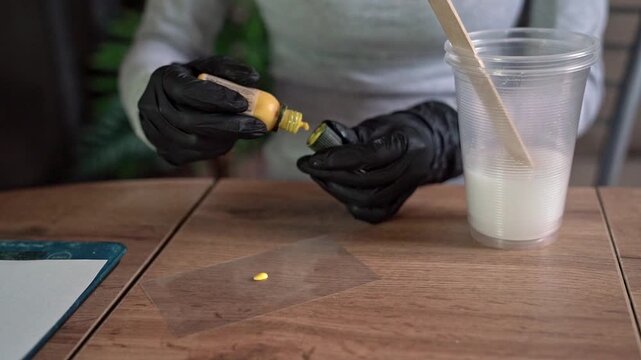 Gloved hands dispensing yellow epoxy pigment drop onto clear acetate beside mixed resin cup in craft workspace with careful mixing tools, focused tabletop setup, creative material preparation