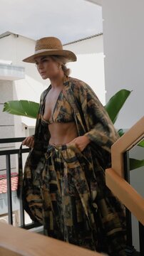 Woman modeling patterned resort wear with bikini top, flowing robe and straw hat in sunlit modern interior.