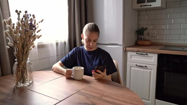 Young woman at kitchen table lowering mug beside smartphone in quiet home interior with soft window light, dried flowers, wooden tabletop, white cabinets, tiled backsplash, and calm domestic mood