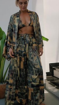 Woman modeling flowing patterned resort wear with bikini top and wide leg pants in bright modern interior.
