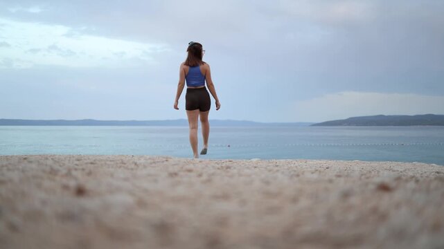 Young woman stretches her quadriceps by calm sea balancing on one leg while holding her foot behind during a peaceful outdoor fitness routine on a quiet summer morning