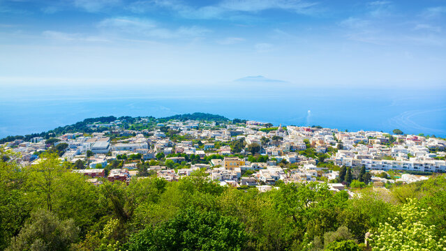 Anacapri panorama stretches above Capri Island, Italy, in Bay of Naples with Mount Vesuvius rising through distant haze. Anacapri stands among green slopes under blue sky beside calm Mediterranean sea
