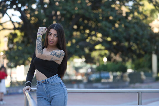 Young Hispanic woman, wearing a black top and jeans, heavily tattooed, with a flirty and seductive look, posing looking at the camera, leaning on a metal railing.