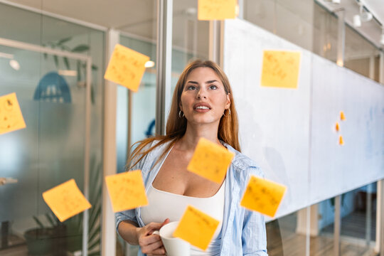 Office brainstorming session concept. Business strategy planning, Woman holding coffee cup and looking at sticky notes on glass wall. Attractive female employee thinking about new ideas.