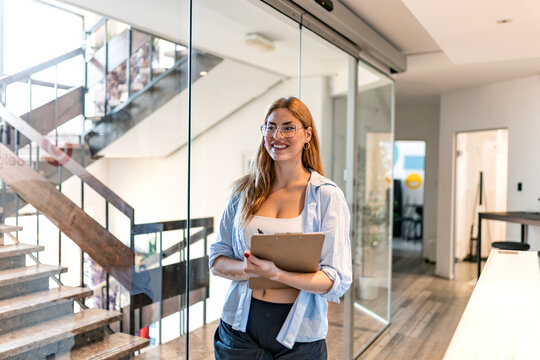 Office corridor portrait of a business woman. Corporate management and administration, Smiling female employee holding clipboard and pen walking in modern office hallway near stairs.