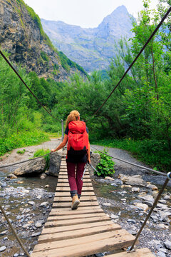 female hiker with red backpack crossing suspension bridge over river in mountain valley, trekking in Alps