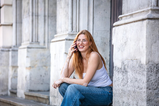 Young woman talking on smartphone sitting on city steps. Mobile communication and urban lifestyle, Smiling female student calling on cell phone near classic stone architecture.