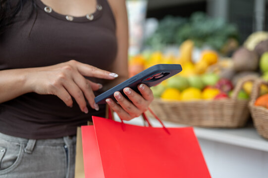 Close up of woman's hands using smartphone and carrying shopping bags while walking in fresh market.