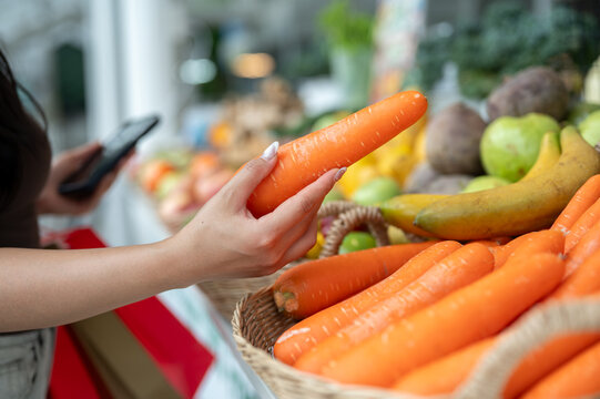 Close up of woman's hand choosing fresh carrots while holding phone and shopping in vegetable market