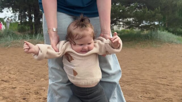 Baby held by caregiver in sandy park smiles beneath beige knitted sweater and sunlit trees, with baby, infant, child, toddler detail, showing kid, caregiver, adult, hand context