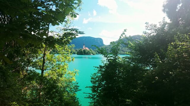 Walking path along the shore of Lake Bled in Slovenia with a scenic view of the iconic Church of the Assumption on Bled Island, framed by trees and green shrubs.
