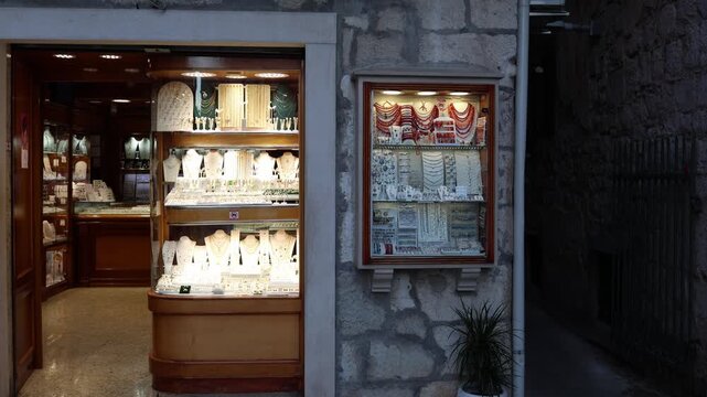 Jewelry shop glows inside an old stone building with necklaces earrings and bracelets displayed in bright windows creating an inviting evening shopping scene for tourists
