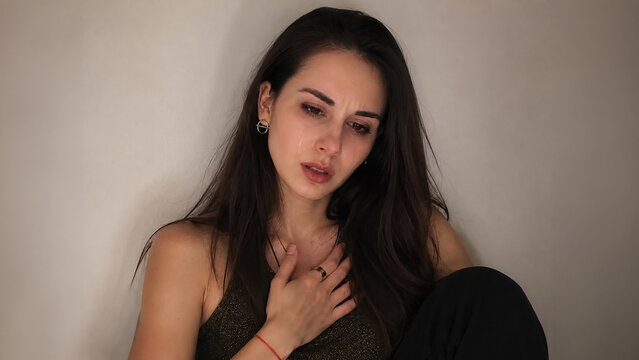 Young woman sitting against a wall with a hand on her chest, experiencing emotional distress. Anxiety, panic attack, stress, loneliness and mental health awareness concept.