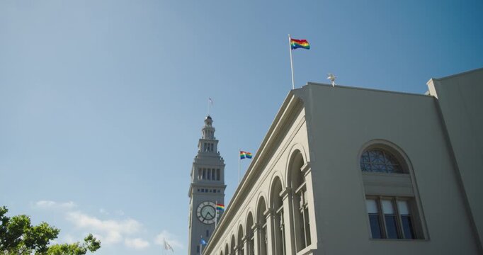 4K cinematic handheld shot of the Ferry Building and clock tower in San Francisco with Pride flags waving and birds flying against a clear blue sky. Iconic waterfront landmark scene.