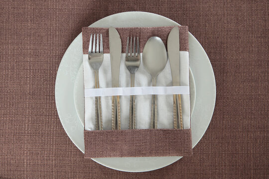 Top view of elegant table setting: cutlery in cloth pocket with white ribbon on white plate on top of brown textured tablecloth