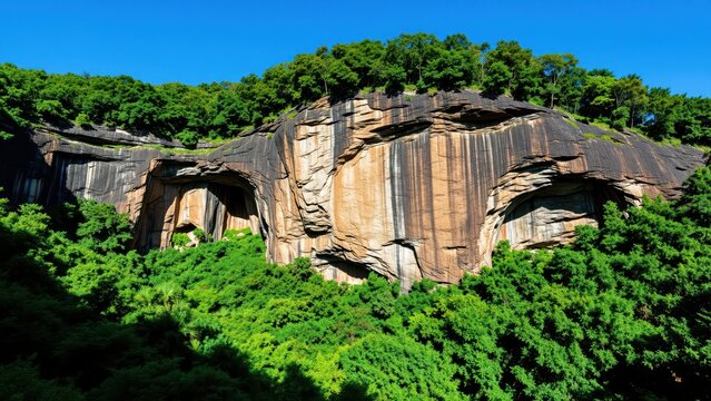 Large rock formation with layered cliffs surrounded by vibrant green trees under a clear blue sky, scenic natural landscape with geological features