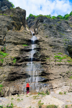 female hiker standing near high waterfall cascading down steep cliff in mountain valley, Alps