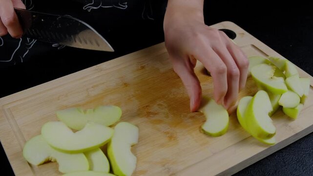 Adult in dark clothing slices green apples with a large knife on a wooden cutting board in a kitchen, Cooking location, medium close-up view.