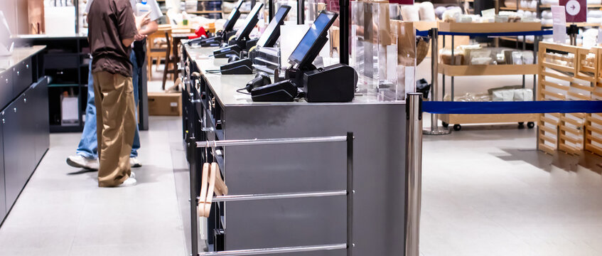 Modern self-checkout counters with digital touch screens and barcode scanners in a retail clothing and home goods department store with customers and blurred background.