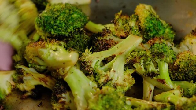Close-up of roasted broccoli florets being stirred with a pink spatula in a pan during cooking in a kitchen, showing browning and seasoning.