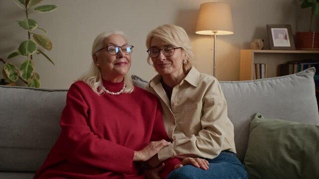 Portrait of Caucasian senior woman and middle aged daughter sitting on sofa, holding hands and smiling at camera. Footage is ideal for senior care services and family support advertisements