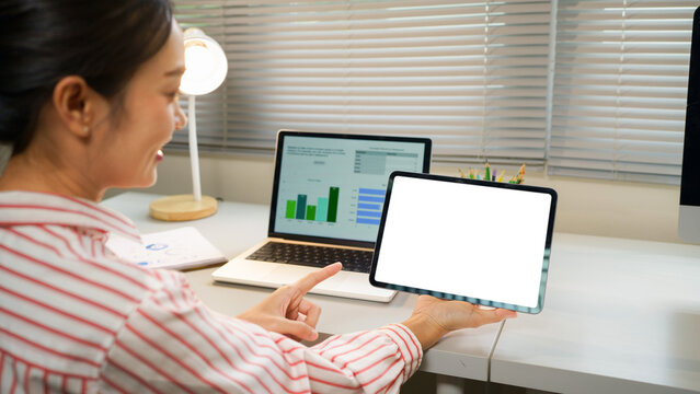 Asian Businesswoman Holding Tablet with Blank White Screen Mockup While Working at Office Desk with Laptop, ideal for app UI and technology presentations.