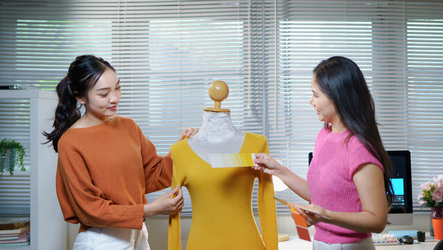 Two young Asian women fashion designers work together with color swatch and outfit sketch works in clothes atelier craft yellow garment on mannequin in sewing workshop creative dressmaker in Studio