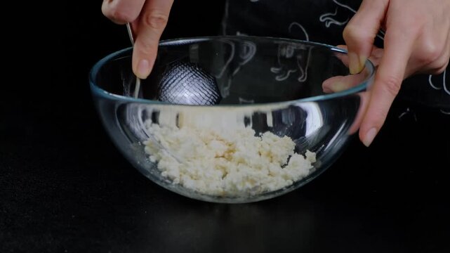 Hands mixing crumbly cheese with a metal strainer in a glass bowl on a dark countertop, cooking preparation in a kitchen, daytime.