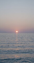 Obraz premium Sun setting over calm ocean horizon with soft pastel sky and reflective water surface, tranquil seascape at dusk conveying peaceful evening mood.