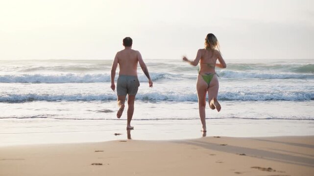 Energetic couple running into sea together at sunset, celebrating freedom and enjoying an active beach lifestyle during summer vacation. Travel, wellness, outdoor recreation and happiness concept