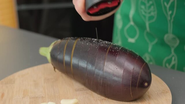 Hand sprinkling white seasoning onto a purple yam on a wooden cutting board in a cooking kitchen, with green patterned cloth and pale yellow food pieces nearby.