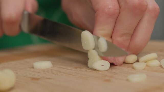 Hands chop white garlic cloves with a silver knife on a wooden cutting board in a Cooking setting, creating piles of minced garlic pieces.