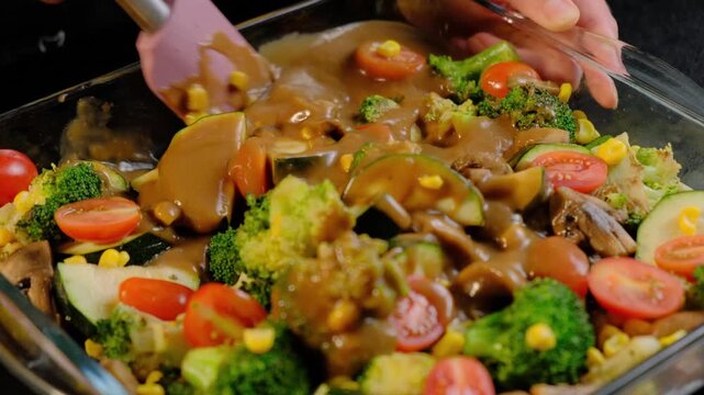 Close-up of hands mixing broccoli, tomatoes, corn, and mushrooms with brown gravy in a glass dish during cooking.