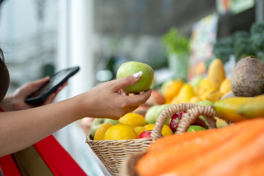 Close up of young woman choosing fresh green apple while using smartphone at fruit market, healthy shopping and grocery retail concept