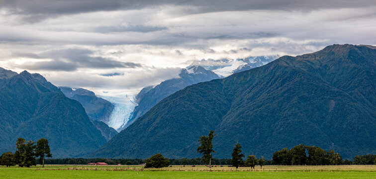 Blick zum Fox Glacier, Suedliche Alpen, Suedinsel, Neuseeland, Gletscherzunge im bewaldeten Tal, gruene Weidelandschaft mit roter Scheune im Vordergrund, dramatischer Wolkenhimmel